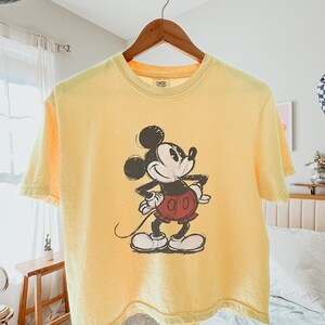 以下が含まれることがあります： ミッキーマウスのイラストが描かれた黄色のTシャツ。漫画のキャラクターは黒、白、赤で描かれています。Tシャツは木製のハンガーに掛けられています。