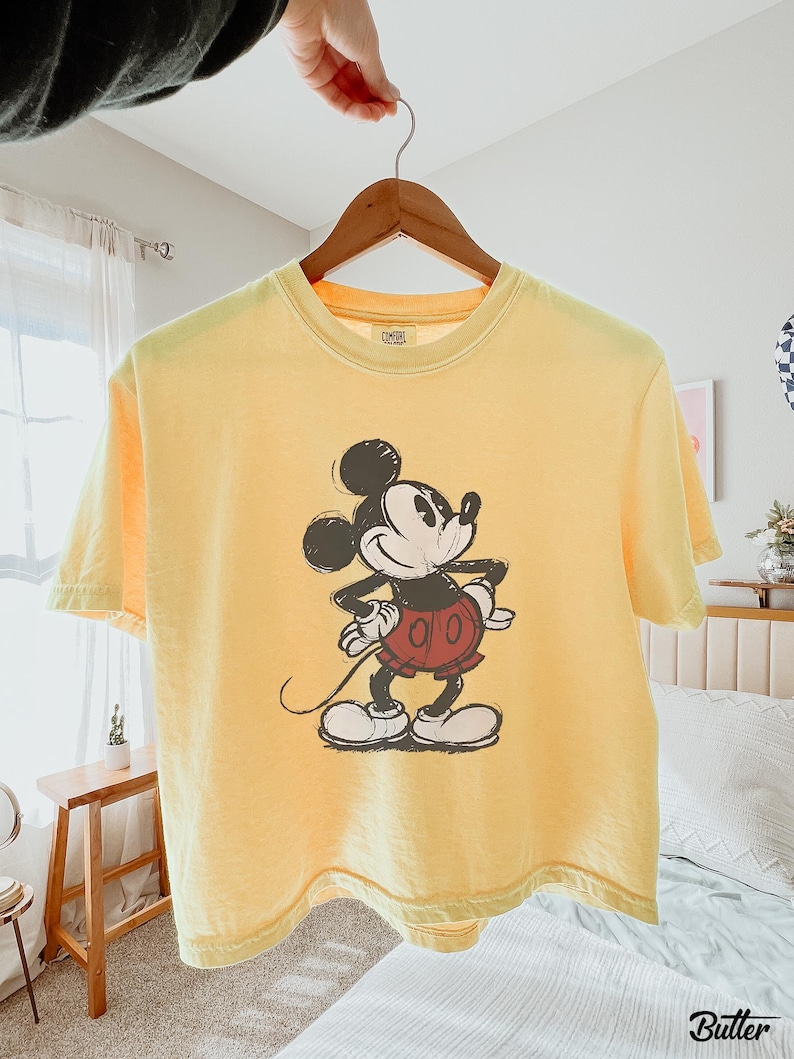 以下が含まれることがあります： ミッキーマウスのイラストが描かれた黄色のTシャツ。漫画のキャラクターは黒、白、赤で描かれています。Tシャツは木製のハンガーに掛けられています。