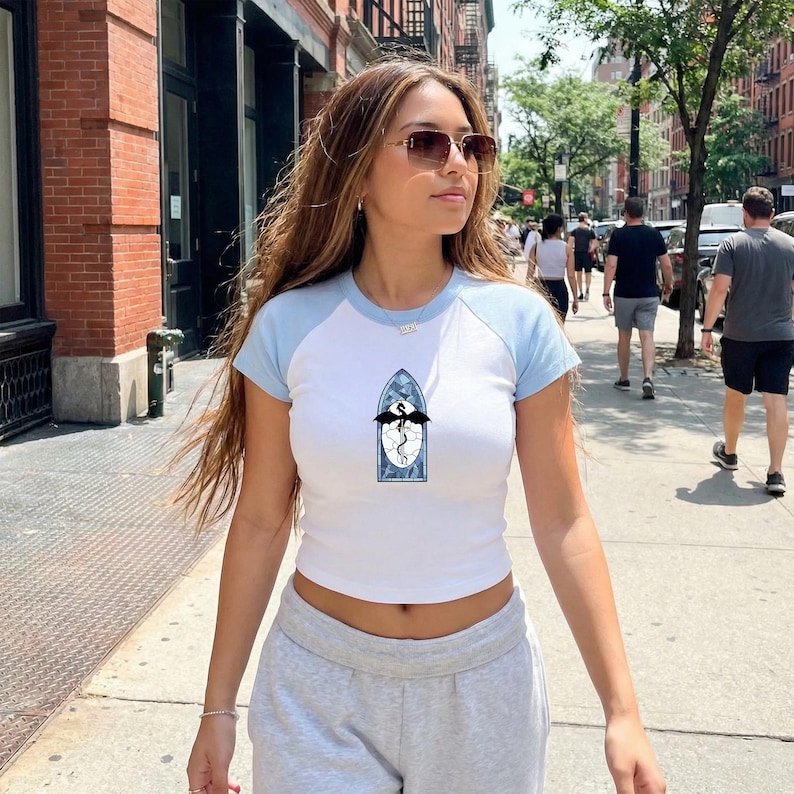 May include: White crop top with light blue sleeves and a crew neck. The shirt features a stained glass design with a black dragon. The model is wearing grey sweatpants and sunglasses.