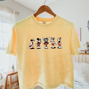 Puede incluir: Una camiseta de manga corta de color amarillo p&aacute;lido con un estampado gr&aacute;fico de cinco figuras de Mickey Mouse en varias poses, cada una con sombrero y chaqueta. La camiseta tiene cuello redondo y est&aacute; colgada en una percha de madera.