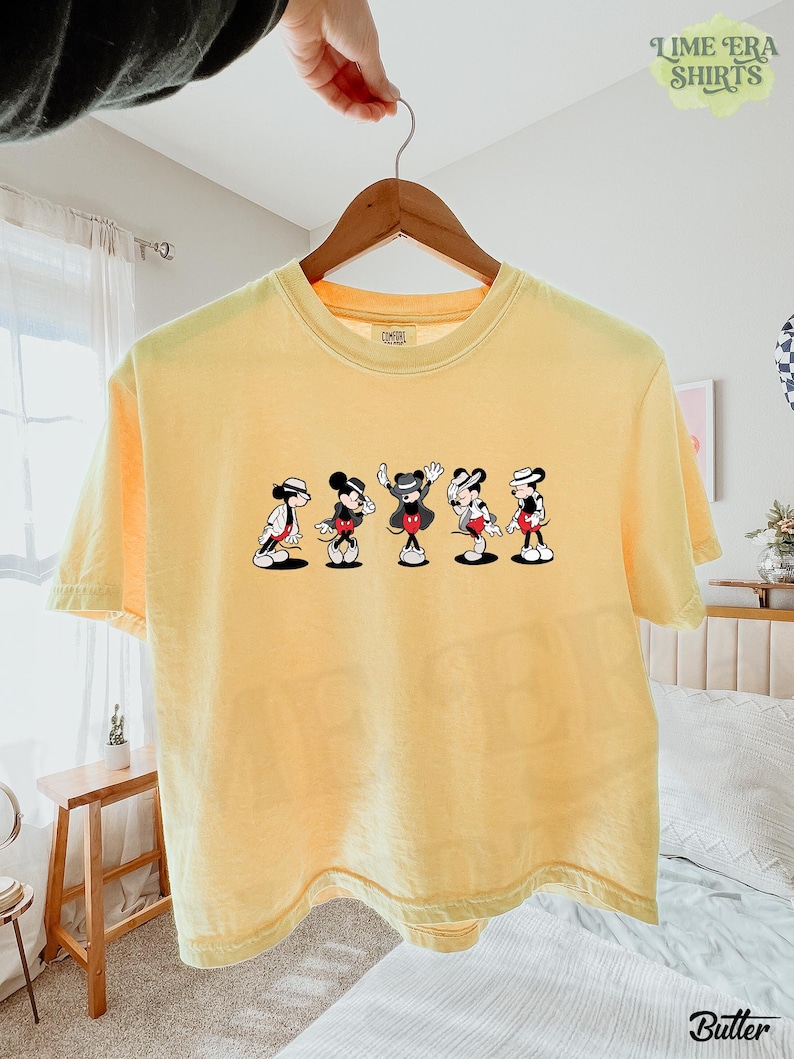 Puede incluir: Una camiseta de manga corta de color amarillo p&aacute;lido con un estampado gr&aacute;fico de cinco figuras de Mickey Mouse en varias poses, cada una con sombrero y chaqueta. La camiseta tiene cuello redondo y est&aacute; colgada en una percha de madera.