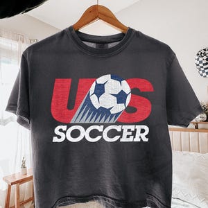 May include: A charcoal gray short-sleeve t-shirt featuring a distressed red graphic of the letters US with a white and blue soccer ball replacing the letter S. The word SOCCER is printed in white block letters underneath the graphic.