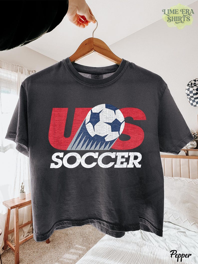 May include: A charcoal gray short-sleeve t-shirt featuring a distressed red graphic of the letters US with a white and blue soccer ball replacing the letter S. The word SOCCER is printed in white block letters underneath the graphic.