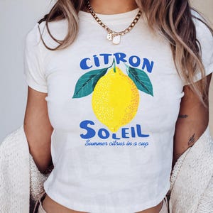 Puede incluir: Una camiseta blanca de manga corta con un gr&aacute;fico de un lim&oacute;n amarillo brillante con hojas verdes. El texto "CITRON SOLEIL" aparece en azul sobre y bajo el lim&oacute;n, con el texto azul m&aacute;s peque&ntilde;o "Summer citrus in a cup" debajo.