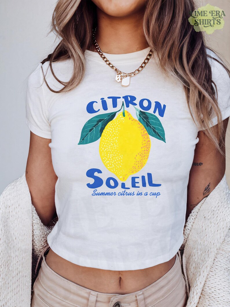 Puede incluir: Una camiseta blanca de manga corta con un gr&aacute;fico de un lim&oacute;n amarillo brillante con hojas verdes. El texto "CITRON SOLEIL" aparece en azul sobre y bajo el lim&oacute;n, con el texto azul m&aacute;s peque&ntilde;o "Summer citrus in a cup" debajo.