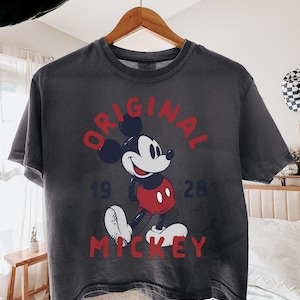 May include: Dark gray t-shirt featuring a classic Mickey Mouse graphic. The design includes the word "ORIGINAL" in red, the year "1928", and the name "MICKEY" in red. The shirt is hanging on a wooden hanger.