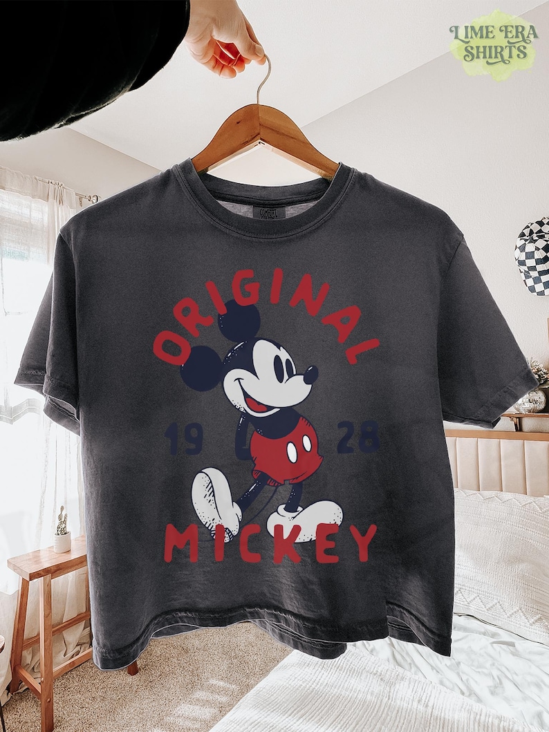 May include: Dark gray t-shirt featuring a classic Mickey Mouse graphic. The design includes the word "ORIGINAL" in red, the year "1928", and the name "MICKEY" in red. The shirt is hanging on a wooden hanger.