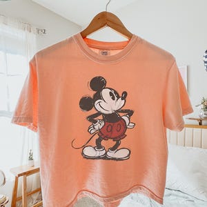 以下が含まれることがあります： スケッチ風のミッキーマウスのグラフィックが特徴のピーチ色のTシャツ。半袖シャツは木製のハンガーに吊るされています。グラフィックは、赤いショートパンツと白い手袋を身に着けた、クラシックな衣装のミッキーマウスを描いています。