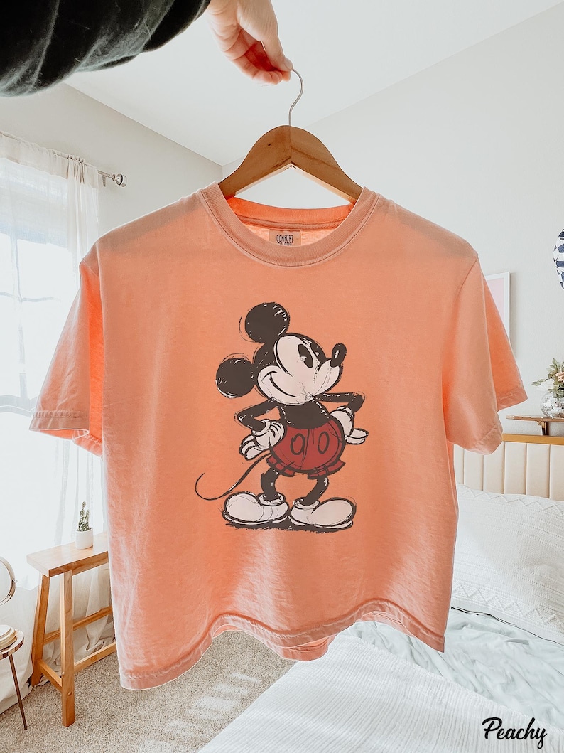 以下が含まれることがあります： スケッチ風のミッキーマウスのグラフィックが特徴のピーチ色のTシャツ。半袖シャツは木製のハンガーに吊るされています。グラフィックは、赤いショートパンツと白い手袋を身に着けた、クラシックな衣装のミッキーマウスを描いています。