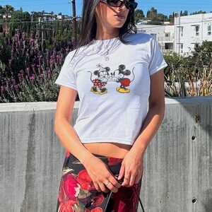 May include: White t-shirt featuring a cartoon image of Mickey and Minnie Mouse kissing. The shirt has a crew neck and short sleeves. The person is wearing a floral print skirt and sunglasses.