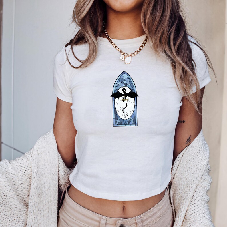 May include: White crop top with a stained glass dragon design. The graphic shows a black dragon with outstretched wings inside a blue and white stained glass window. The shirt is short-sleeved and has a round neckline.