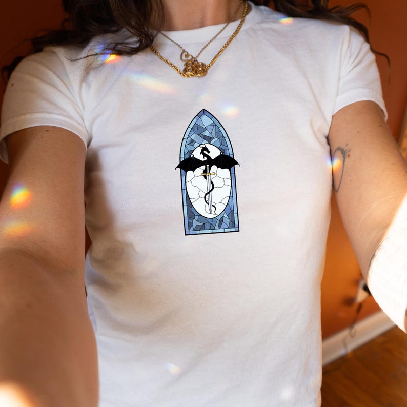 May include: White t-shirt with a stained-glass style design. The design features a black dragon, sword, and wings, set within a blue and white frame. The shirt is made of a soft, white material.