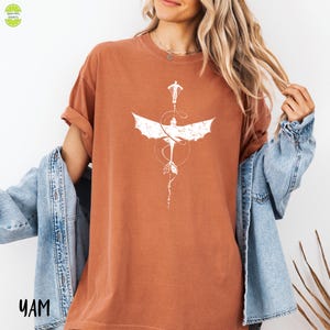 May include: A rust-coloured t-shirt with a white graphic of a dragon with outstretched wings and a person. The design includes a dotted circle and arrow details. The shirt has short sleeves and a relaxed fit.