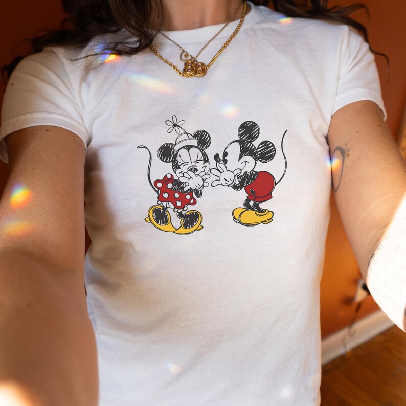 May include: White t-shirt featuring a black and red graphic of Mickey and Minnie Mouse. Minnie is wearing a red polka dot dress and Mickey is wearing red shorts. Both characters are smiling and interacting.