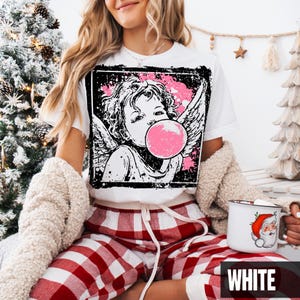 May include: White t-shirt featuring a black and pink graphic of an angel blowing a bubblegum bubble. The angel has wings and is set against a distressed background. The model is wearing red and white plaid pajama pants.