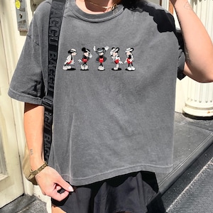 Puede incluir: Una camiseta de manga corta en color gris carb&oacute;n con un estampado gr&aacute;fico de cinco figuras de Mickey Mouse en varias poses, cada una con sombrero y pantalones cortos rojos. La camiseta tiene cuello redondo y un corte holgado.