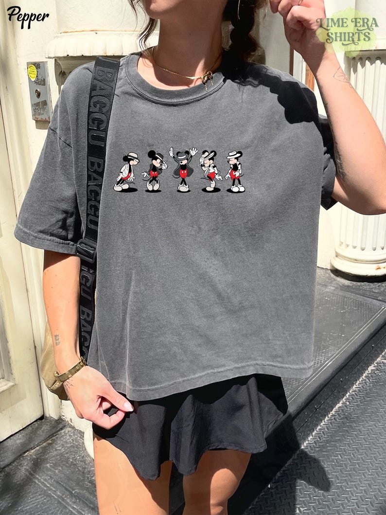 Puede incluir: Una camiseta de manga corta en color gris carb&oacute;n con un estampado gr&aacute;fico de cinco figuras de Mickey Mouse en varias poses, cada una con sombrero y pantalones cortos rojos. La camiseta tiene cuello redondo y un corte holgado.