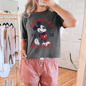 May include: Dark gray t-shirt with a vintage Mickey Mouse graphic, paired with red and white checkered shorts. The shirt features the text "ORIGINAL MICKEY 1928" in a distressed style. The shorts have a classic button-fly design.