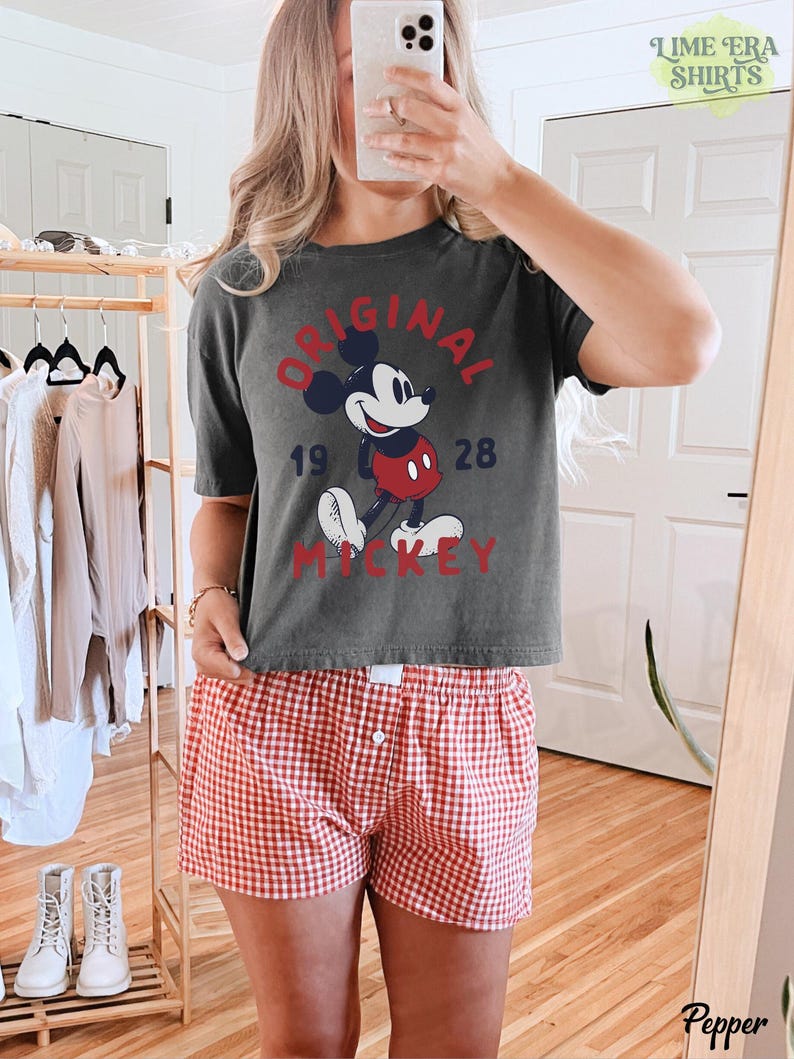 May include: Dark gray t-shirt with a vintage Mickey Mouse graphic, paired with red and white checkered shorts. The shirt features the text "ORIGINAL MICKEY 1928" in a distressed style. The shorts have a classic button-fly design.