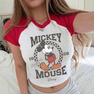 May include: White crop top with red sleeves featuring a vintage Mickey Mouse graphic. The graphic includes the words "Mickey Mouse" and "Disney" with the year "1928". The shirt has a classic baseball tee design.