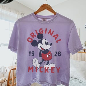 May include: A lavender t-shirt featuring a graphic of Mickey Mouse with the text "ORIGINAL 1928 MICKEY". The shirt is hanging on a wooden hanger. The brand name "TIME ERA SHIRTS" is visible.