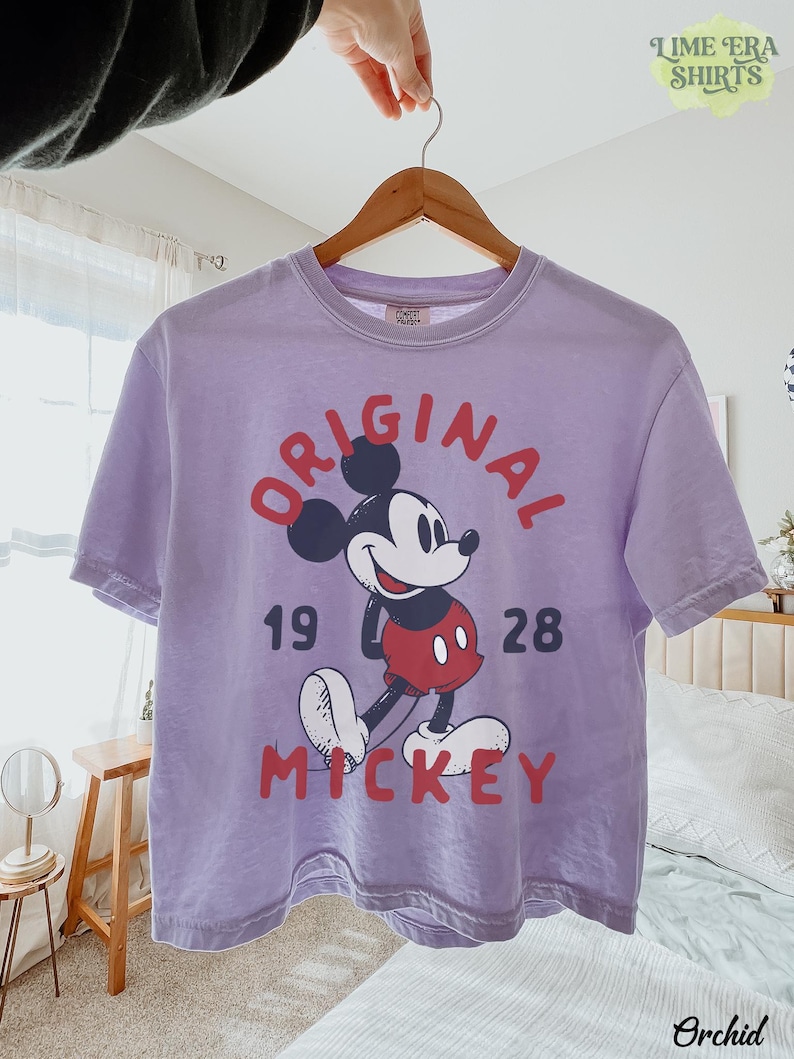 May include: A lavender t-shirt featuring a graphic of Mickey Mouse with the text "ORIGINAL 1928 MICKEY". The shirt is hanging on a wooden hanger. The brand name "TIME ERA SHIRTS" is visible.