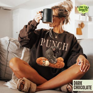 May include: Dark chocolate-colored sweatshirt with the word "PUNCH" printed across the chest. The sweatshirt features a graphic of a small monkey sitting on a stuffed animal. A black mug is held up.
