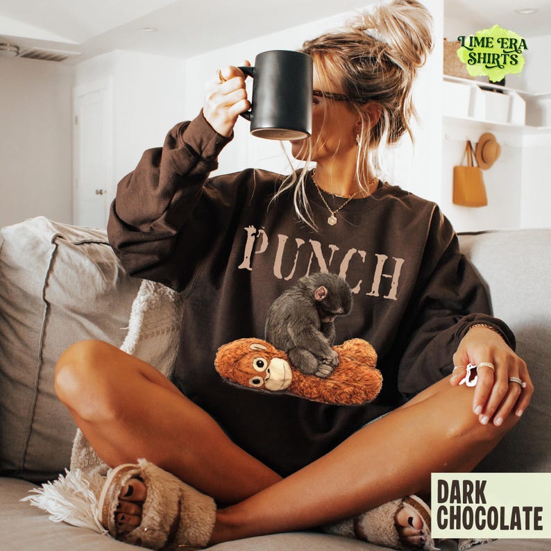 May include: Dark chocolate-colored sweatshirt with the word "PUNCH" printed across the chest. The sweatshirt features a graphic of a small monkey sitting on a stuffed animal. A black mug is held up.