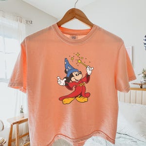 May include: Peach-colored t-shirt featuring a graphic of Mickey Mouse dressed as a wizard, holding a wand with stars. The shirt is hanging on a wooden hanger. The word "Peachy" is printed in the bottom right corner.