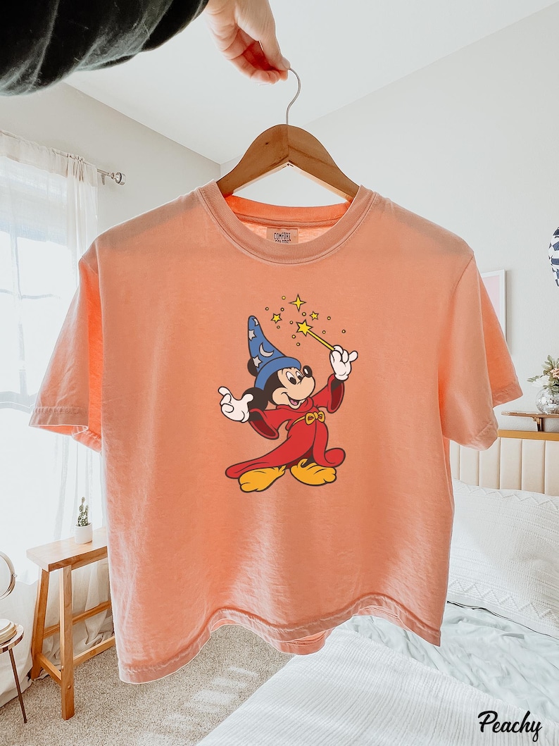 May include: Peach-colored t-shirt featuring a graphic of Mickey Mouse dressed as a wizard, holding a wand with stars. The shirt is hanging on a wooden hanger. The word "Peachy" is printed in the bottom right corner.