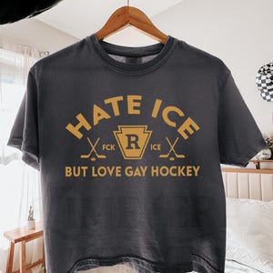 May include: Dark gray t-shirt with the text "HATE ICE BUT LOVE GAY HOCKEY" in gold lettering. The design includes crossed hockey sticks and a gold emblem. The shirt is hanging on a wooden hanger.