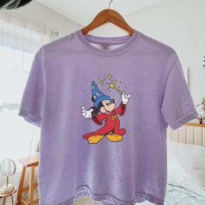 May include: A light purple t-shirt featuring a cartoon Mickey Mouse dressed as a wizard, holding a wand with sparkling stars. The shirt has a crew neck and short sleeves. The word "Orchid" is printed on the bottom right.
