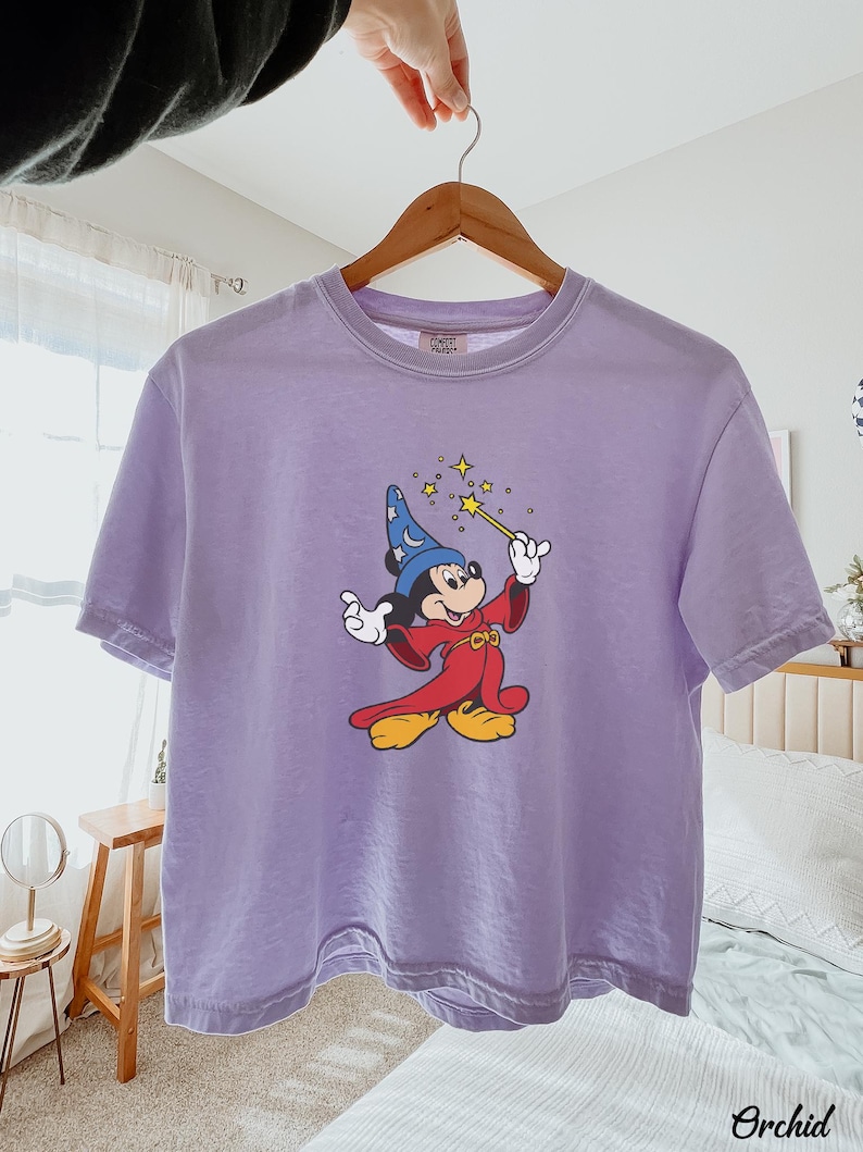 May include: A light purple t-shirt featuring a cartoon Mickey Mouse dressed as a wizard, holding a wand with sparkling stars. The shirt has a crew neck and short sleeves. The word "Orchid" is printed on the bottom right.