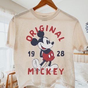 May include: Ivory-colored t-shirt featuring a graphic of Mickey Mouse with the words "ORIGINAL MICKEY 1928" in red. The shirt is hanging on a wooden hanger. The brand name "Lime Era Shirts" is visible in the top right corner.