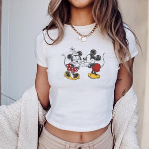 May include: White crop top featuring a black and red graphic of Mickey and Minnie Mouse. The cartoon characters are depicted in a classic pose, with Minnie wearing a polka dot dress and Mickey in red shorts. The shirt has a crew neck and short sleeves.