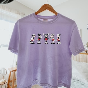 Puede incluir: Una camiseta de manga corta de color morado claro con un estampado gr&aacute;fico de cinco ratones de dibujos animados en varias poses de baile, cada uno con sombrero y chaqueta. La camiseta tiene cuello redondo y cuelga de una percha de madera.
