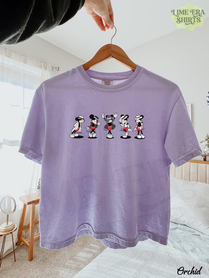 Puede incluir: Una camiseta de manga corta de color morado claro con un estampado gr&aacute;fico de cinco ratones de dibujos animados en varias poses de baile, cada uno con sombrero y chaqueta. La camiseta tiene cuello redondo y cuelga de una percha de madera.