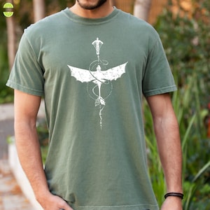 May include: Sage green t-shirt with a white dragon graphic. The design includes a person at the top, wings, a dotted circle, and a flowing tail. The shirt has short sleeves and a crew neck. The t-shirt is a muted green colour.