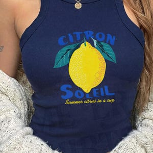 Puede incluir: Una camiseta de tirantes acanalada de color azul marino con un gr&aacute;fico de un lim&oacute;n amarillo con hojas verdes. El texto CITRON SOLEIL aparece en letras azules sobre y bajo el lim&oacute;n, con la frase amarilla Summer citrus in a cup debajo.