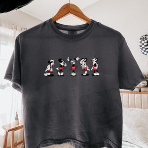 Puede incluir: Una camiseta de manga corta en color gris carb&oacute;n con un estampado gr&aacute;fico de cinco figuras de Mickey Mouse en varias poses, cada una con sombrero y chaqueta. La camiseta tiene cuello redondo y un corte relajado sobre una percha de madera.