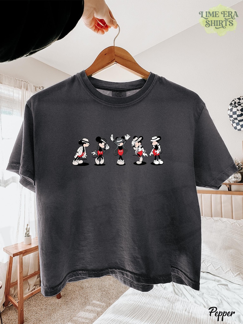 Puede incluir: Una camiseta de manga corta en color gris carb&oacute;n con un estampado gr&aacute;fico de cinco figuras de Mickey Mouse en varias poses, cada una con sombrero y chaqueta. La camiseta tiene cuello redondo y un corte relajado sobre una percha de madera.