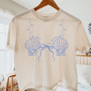 May include: Ivory-colored t-shirt with a blue seashell design and the text "Jennifer's Bachelorette" above the shells. Below the shells are the words "Miami, FL" and "June 2024". The shirt is hanging on a wooden hanger.