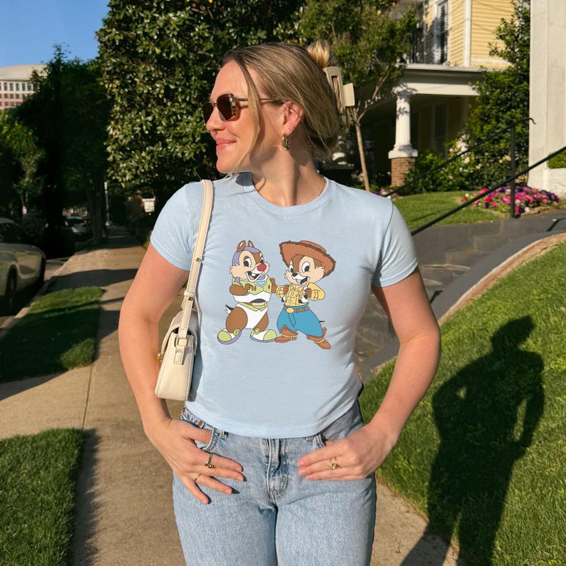 May include: Light blue t-shirt featuring a cartoon graphic of Chip and Dale. The shirt has short sleeves and a crew neck. The graphic shows Chip dressed as a baby and Dale as a cowboy. The shirt is paired with light wash denim jeans.