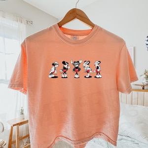 Puede incluir: Una camiseta de manga corta color melocot&oacute;n con un estampado gr&aacute;fico de cinco figuras de Mickey Mouse en varias poses de baile, cada una con un sombrero fedora y un traje. La camiseta tiene cuello redondo y cuelga de una percha de madera.