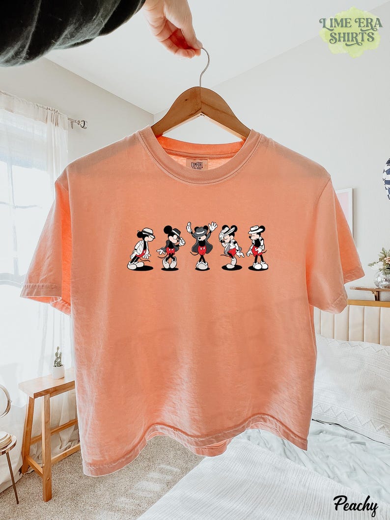 Puede incluir: Una camiseta de manga corta color melocot&oacute;n con un estampado gr&aacute;fico de cinco figuras de Mickey Mouse en varias poses de baile, cada una con un sombrero fedora y un traje. La camiseta tiene cuello redondo y cuelga de una percha de madera.