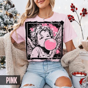 May include: Pink t-shirt featuring a graphic of a cherub blowing a bubblegum bubble. The cherub is in a black frame with pink splatter accents. The word "PINK" is printed at the bottom of the shirt.