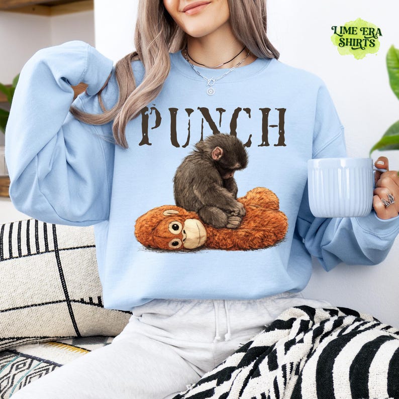May include: Light blue sweatshirt with the word "PUNCH" in brown letters. The sweatshirt features a graphic of a monkey sitting on a stuffed animal. The sweatshirt has long sleeves and a crew neck.
