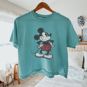 以下が含まれることがあります： ミッキーマウスのヴィンテージプリントが施された、ティールカラーのTシャツ。漫画のキャラクターは黒、白、赤で描かれています。Tシャツは木製のハンガーに掛けられています。カジュアルウェアです。
