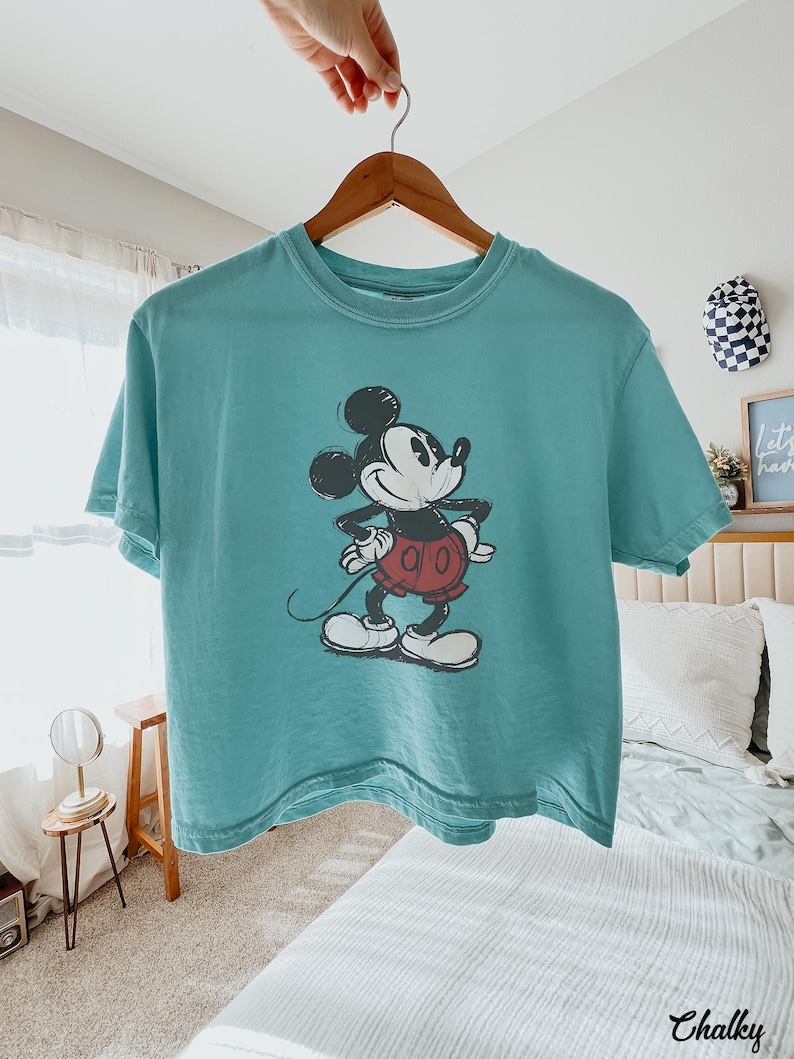 以下が含まれることがあります： ミッキーマウスのヴィンテージプリントが施された、ティールカラーのTシャツ。漫画のキャラクターは黒、白、赤で描かれています。Tシャツは木製のハンガーに掛けられています。カジュアルウェアです。