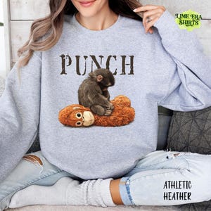 May include: Athletic heather gray sweatshirt with the word "PUNCH" in brown letters. The graphic features a brown monkey sitting on a brown teddy bear. The sweatshirt is made of a soft material.